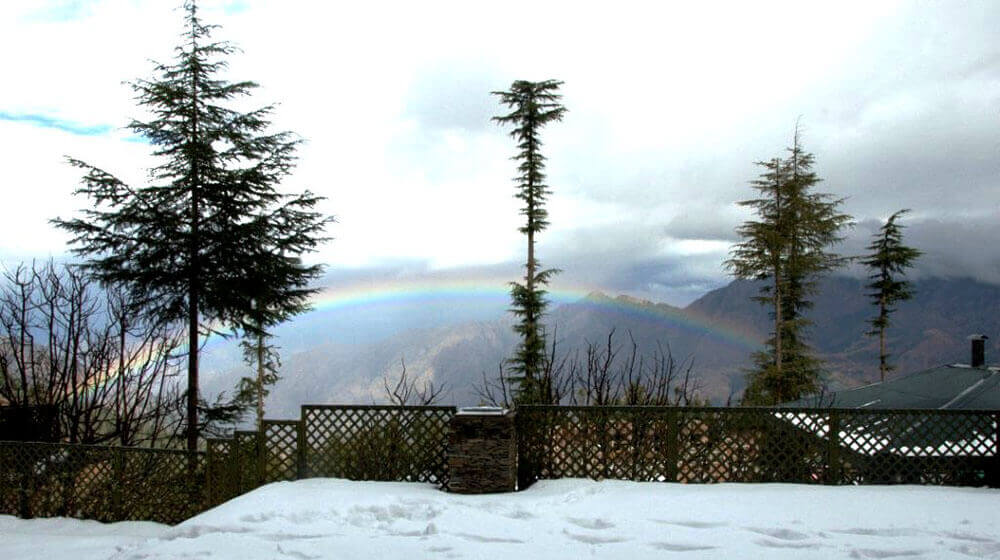A two-story mountain villa with warm lights glowing from the windows and decorative string lights on the balcony, surrounded by a green lawn, blooming hydrangeas, and white patio furniture.