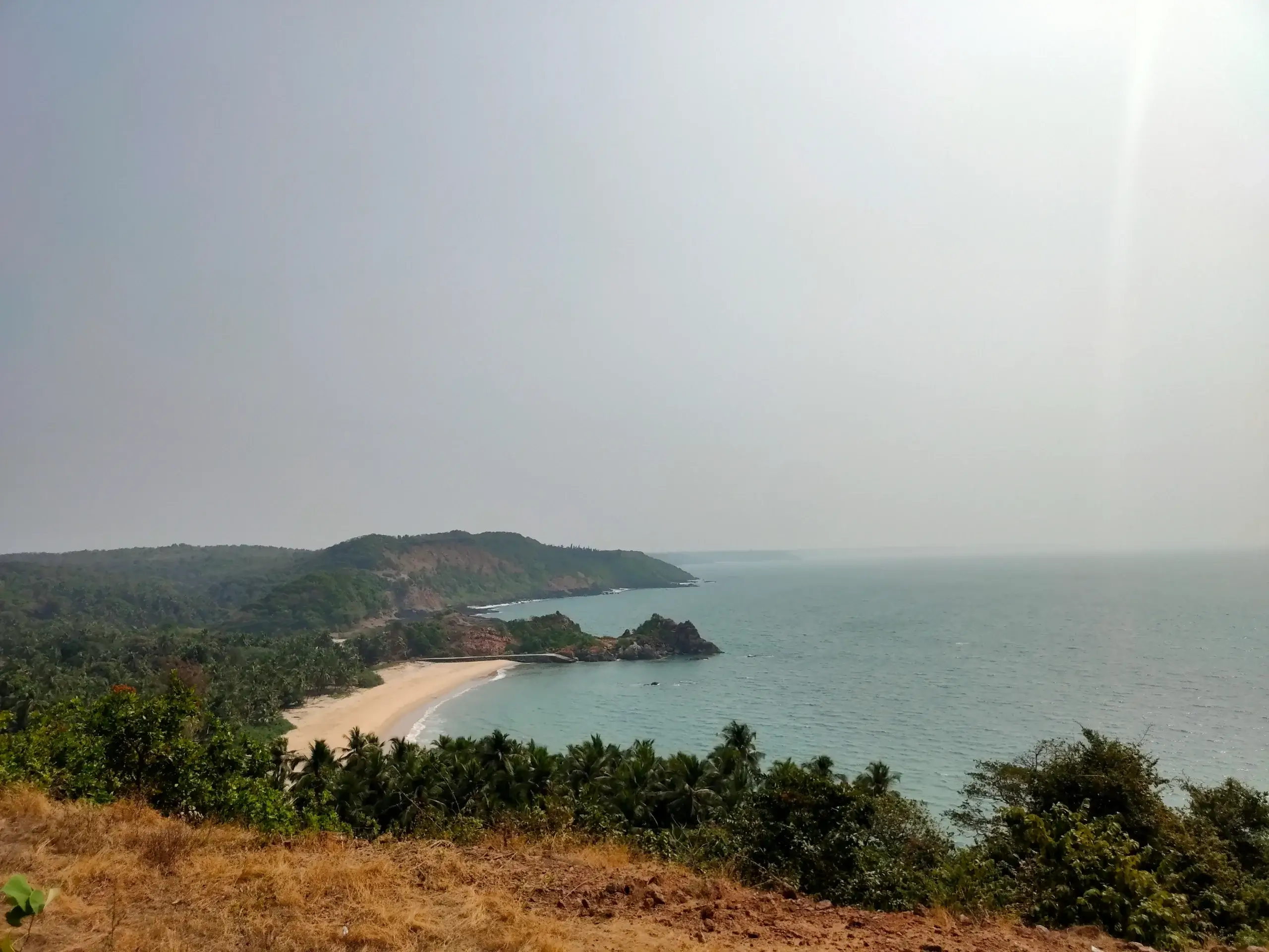 A scenic coastal view of a secluded beach with turquoise waters, palm trees, and rocky cliffs along the Konkan coastline.