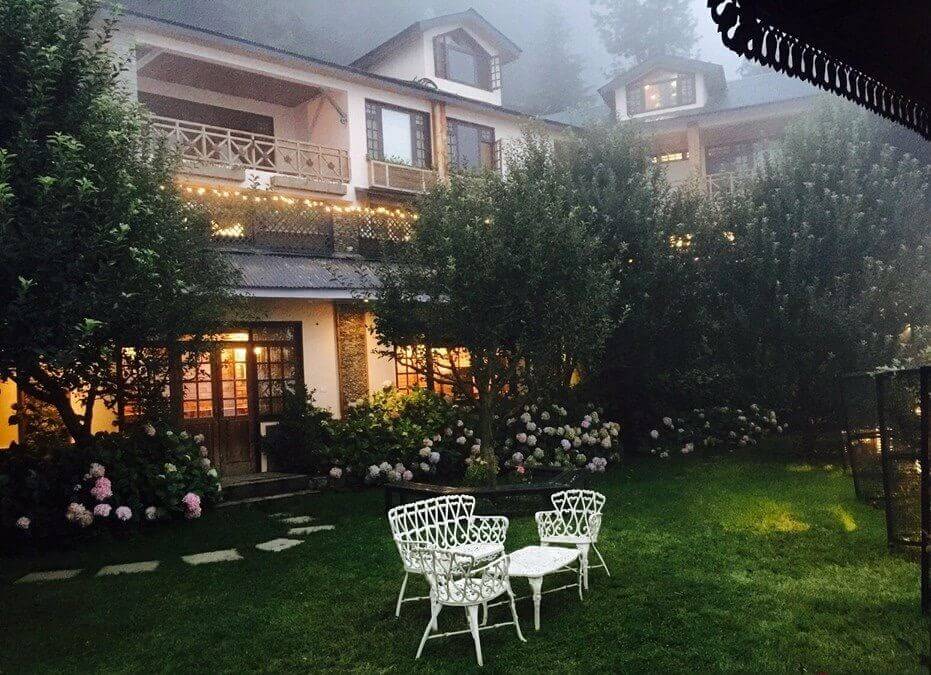 A cozy, mountain villa with warm yellow lights at dusk, featuring a green lawn with white wrought iron garden chairs and tables, surrounded by hydrangea flowers and misty pine trees in the background.