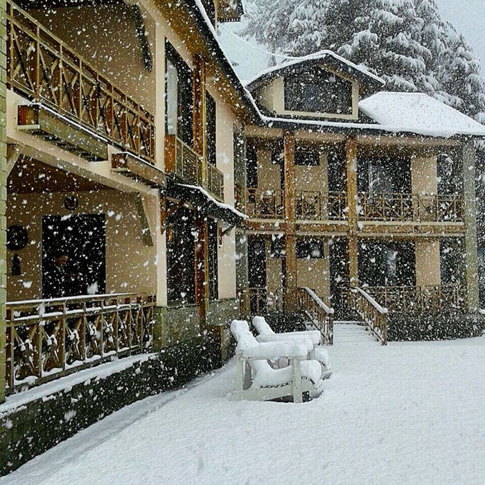 A close-up view of the same mountain villa during a heavy snowstorm, with thick snowflakes filling the air and white lounge chairs completely covered in a layer of fresh snow.