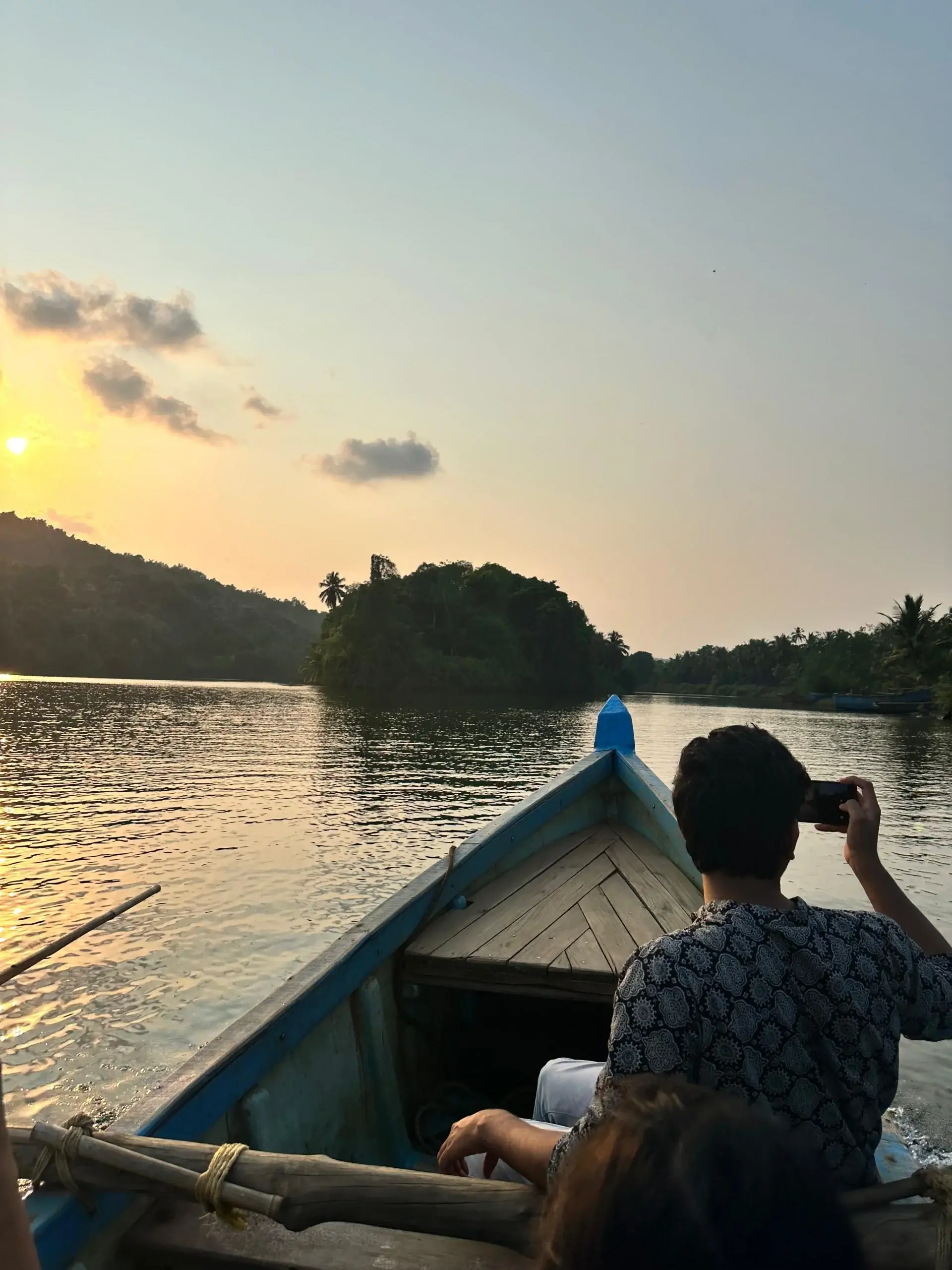 A traveller seated on a wooden boat cruising through calm Konkan backwaters during sunset, with palm-covered hills and golden reflections on the water.