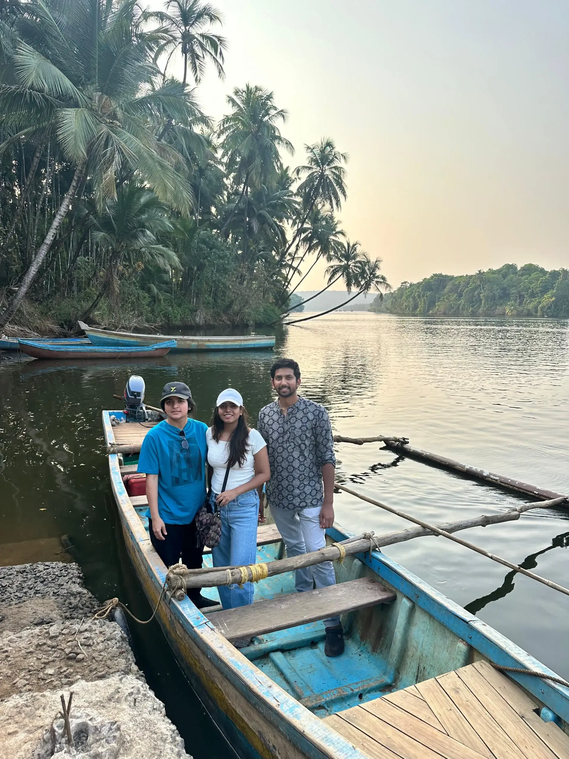 Three travellers standing on a traditional wooden boat by a calm river, surrounded by coconut palms and lush greenery in coastal Konkan.