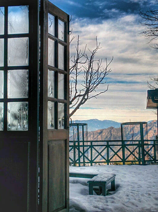 A high-angle view of a villa’s terrace covered in deep snow under a bright blue sky with white clouds, overlooking a valley of evergreen trees and distant mountains.